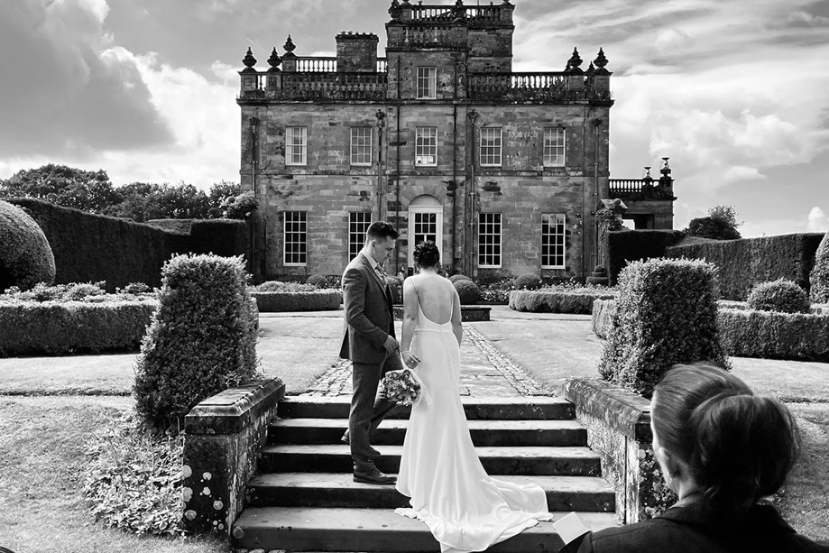 Black and white image of bride and groom walking up steps in front of country house venue